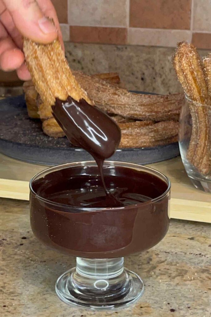 A homemade churro dipped into rich dark chocolate sauce with freshly made churros in the background.