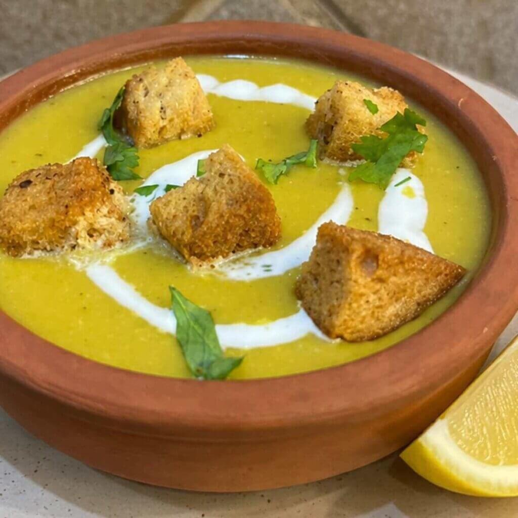 Creamy yellow lentil soup served in a rustic clay bowl on a ceramic plate, topped with crunchy homemade croutons, a swirl of cream, fresh herbs, and a wedge of lemon on the side.