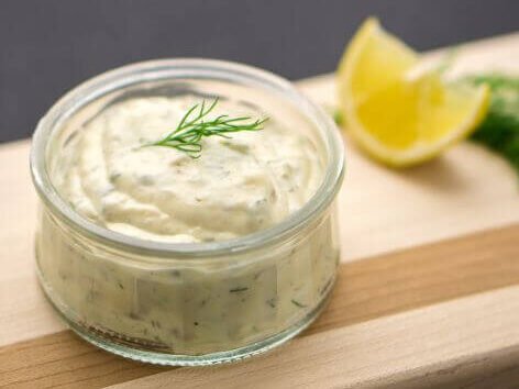 Creamy homemade tartar sauce in a glass jar garnished with fresh dill, served with lemon wedges on a wooden board.