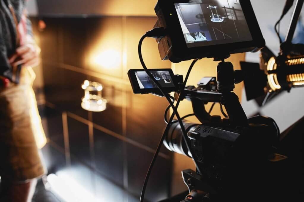 Professional video camera filming a cooking tutorial in a modern kitchen studio with lighting equipment and a blurred presenter in the background.