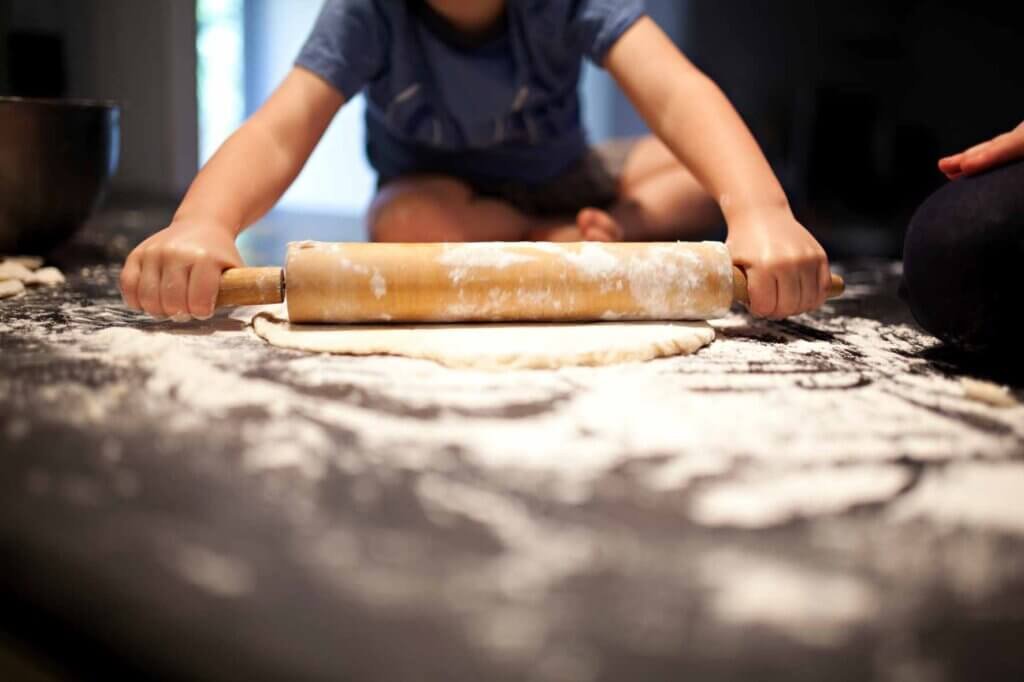 Child using a wooden rolling pin to flatten homemade dough on a floured kitchen countertop while learning to bake.