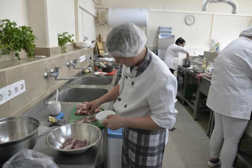Culinary student preparing ingredients in a professional training kitchen while classmates cook in the background.