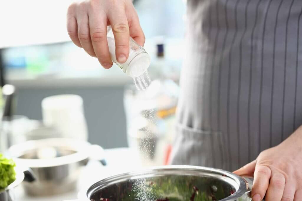 Chef adding salt gradually to a saucepan while seasoning a sauce, demonstrating the importance of tasting and seasoning in stages.