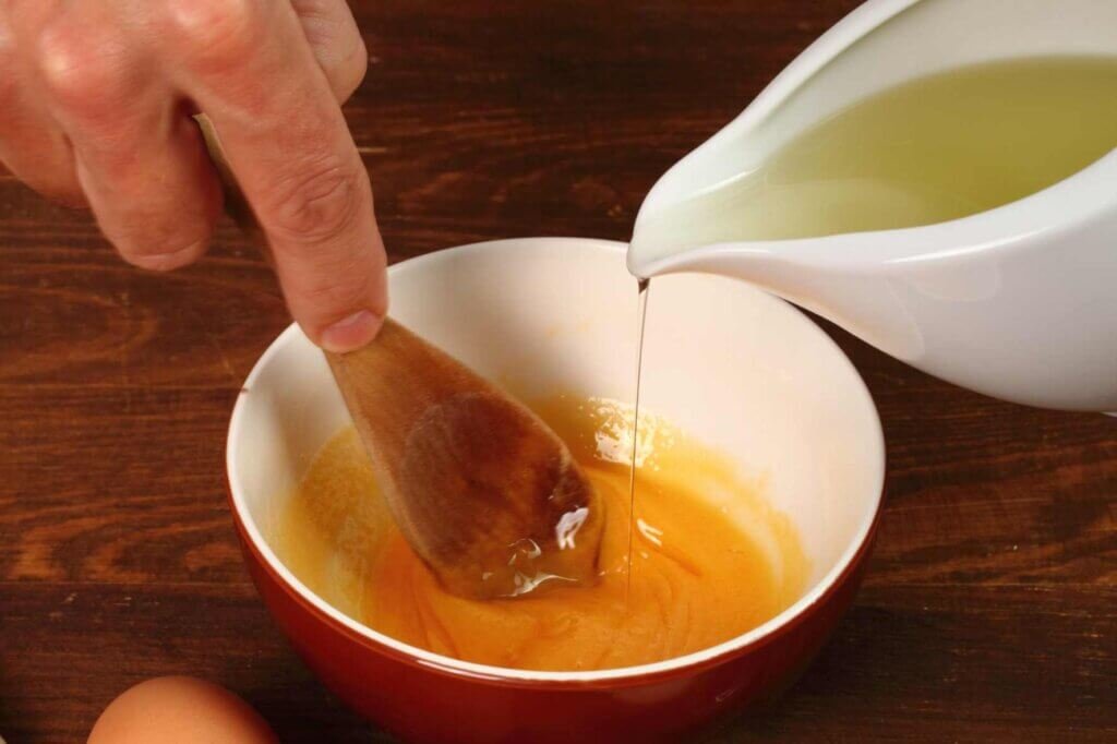 Oil being slowly poured into egg yolks while stirring to create a stable emulsion for homemade mayonnaise.