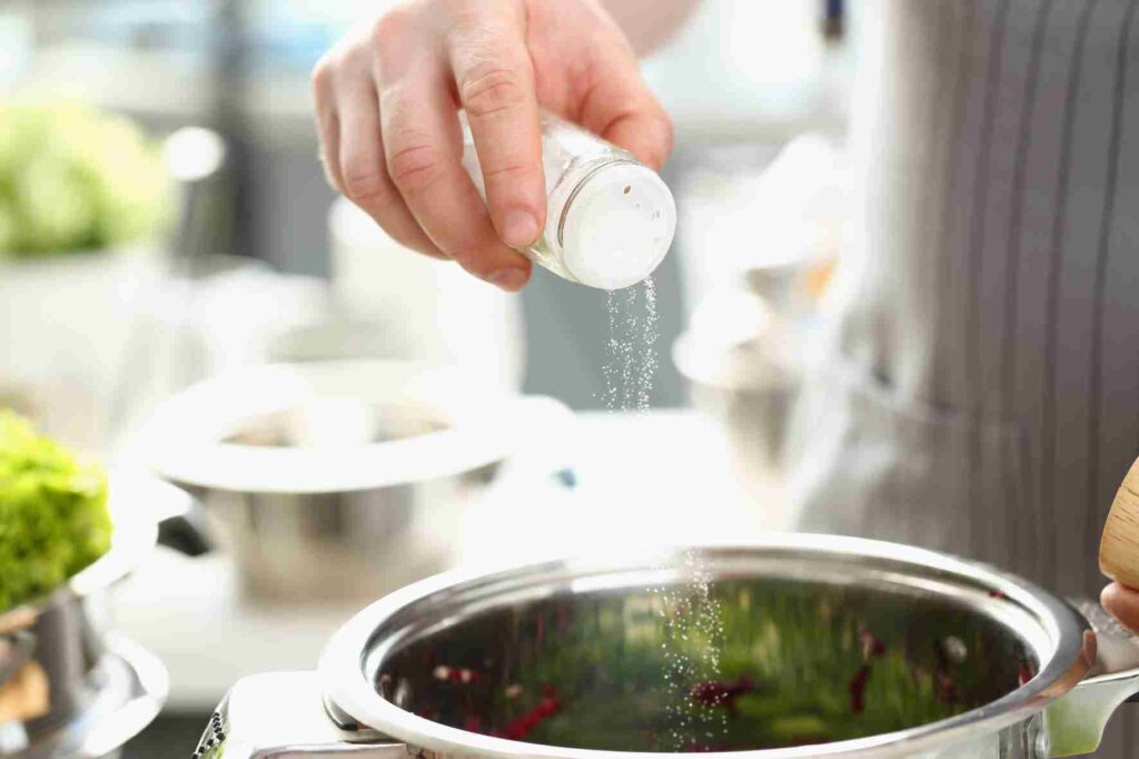 A chef sprinkling salt from a shaker into a pot while cooking in a professional kitchen.