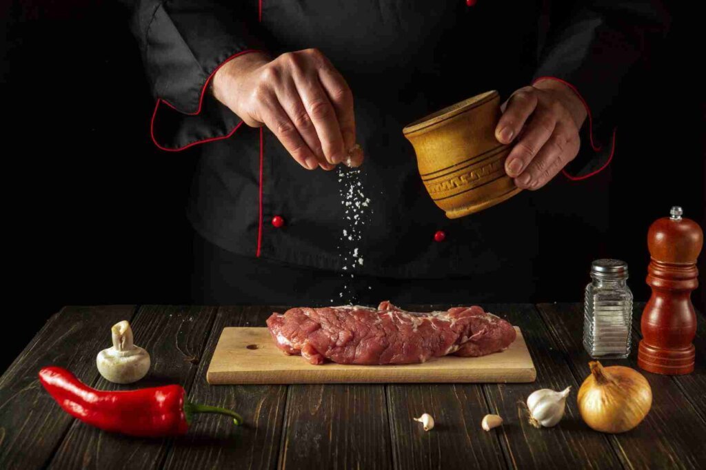 A chef sprinkling coarse salt over raw meat on a wooden cutting board before cooking.