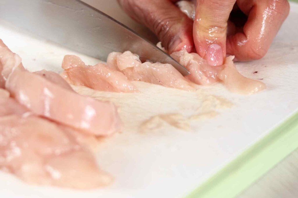 Chef cutting raw chicken on a plastic cutting board with a kitchen knife.