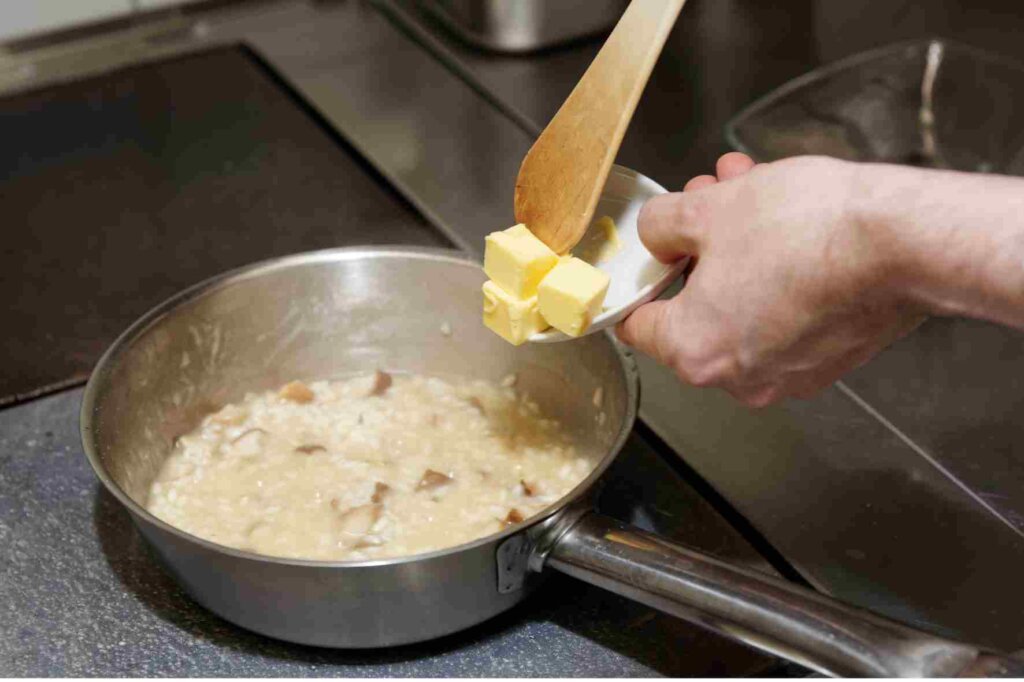 A chef adding cold butter cubes to a creamy mushroom sauce in a saucepan.