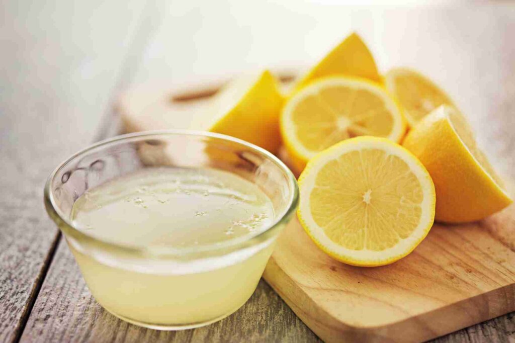 Fresh lemon juice in a glass bowl beside halved lemons and lemon wedges on a wooden cutting board.