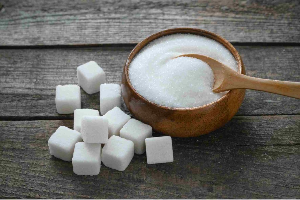 White granulated sugar in a wooden bowl with a spoon and sugar cubes on a rustic table.