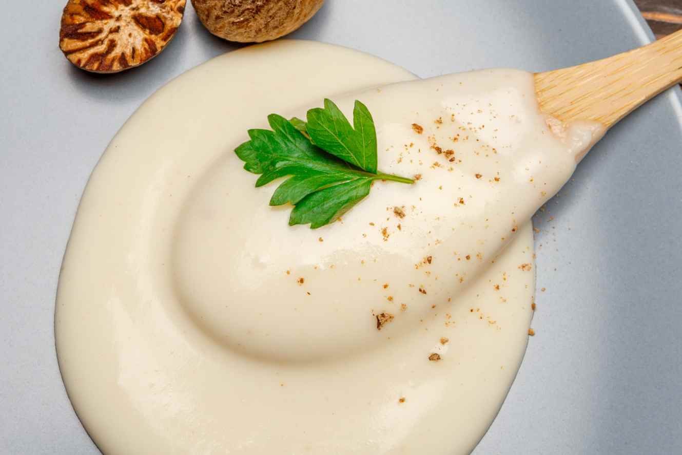 Smooth and creamy homemade béchamel sauce coating a spatula with nutmeg and parsley garnish.