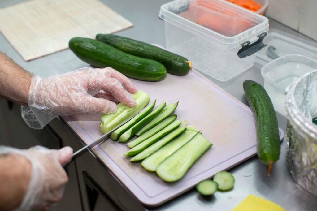 A cook wearing gloves slices fresh cucumbers lengthwise on a cutting board.