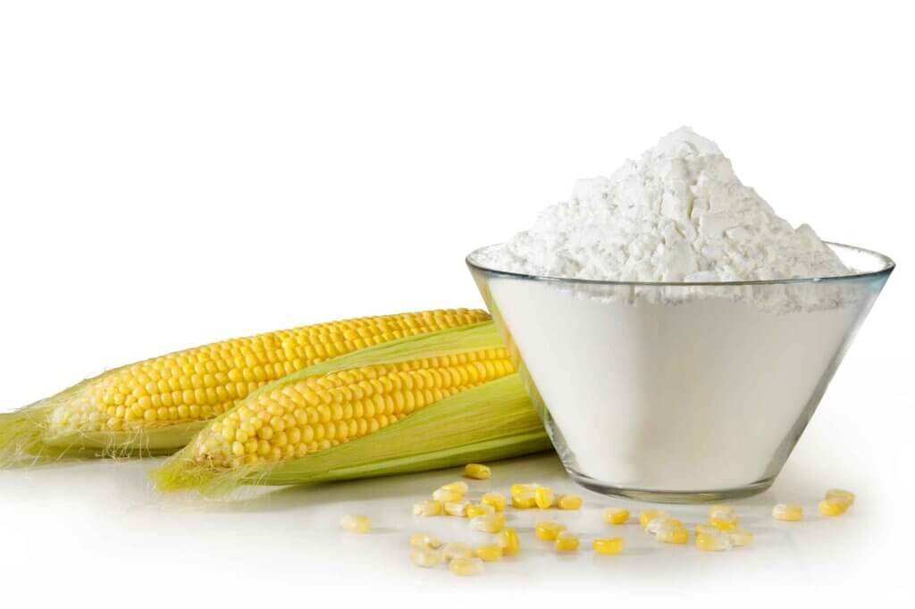 A glass bowl filled with cornstarch beside fresh yellow corn on a white background