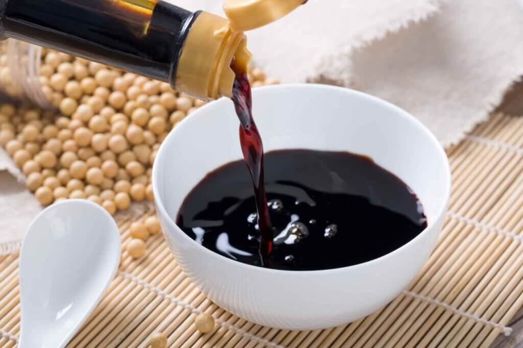 Soy sauce being poured into a white bowl beside dried soybean