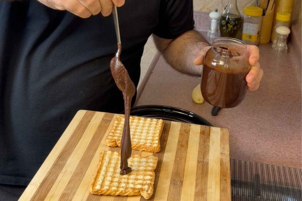 Thick homemade hazelnut chocolate spread drizzled over toasted bread while a jar of the spread is held nearby.