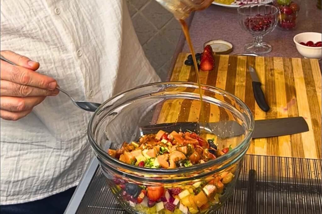 Chef François pouring a golden honey-based dressing over freekeh salad with chopped fruits, vegetables, and pomegranate seeds in a glass mixing bowl.