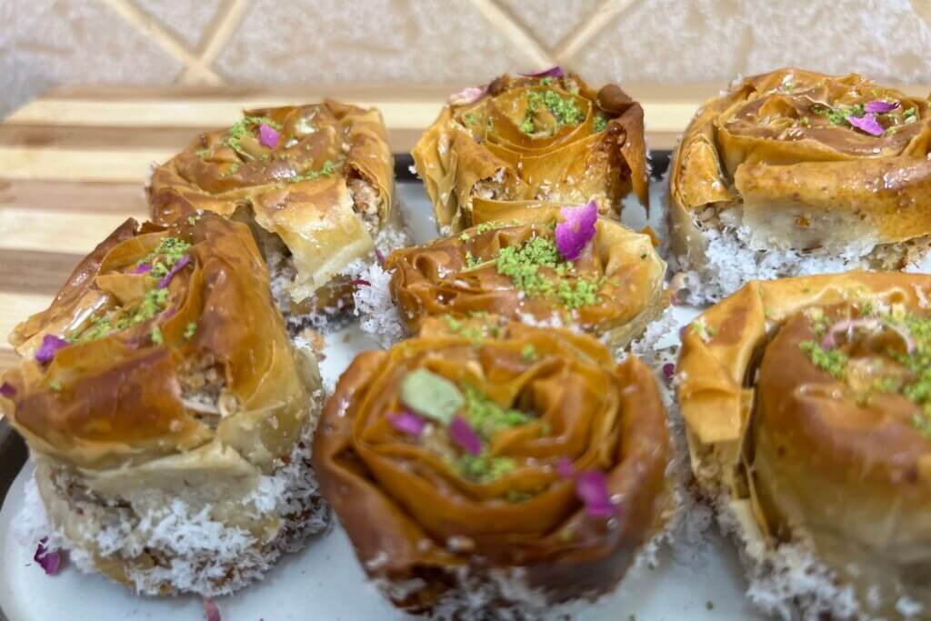Close-up of golden baklava roses filled with almonds and coconut, topped with crushed pistachios and rose petals on a white plate