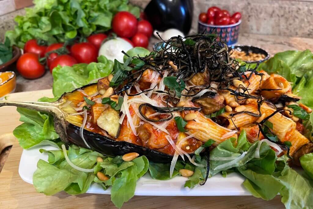 an eggplant boat stuffed with rigatoni alla Siciliana, place in a white dish decorated with lettuce. Behind the dish a display of tomatoes, eggplants, and lettuce.