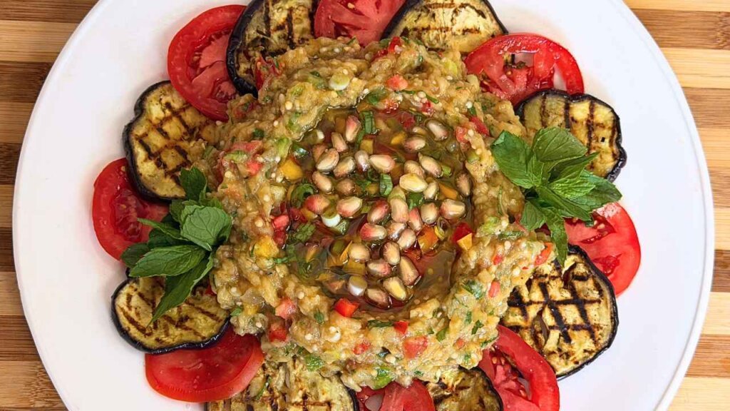 Overhead view of Eggplant Raheb salad with pomegranate seeds and mint, surrounded by alternating grilled eggplant and tomato slices.