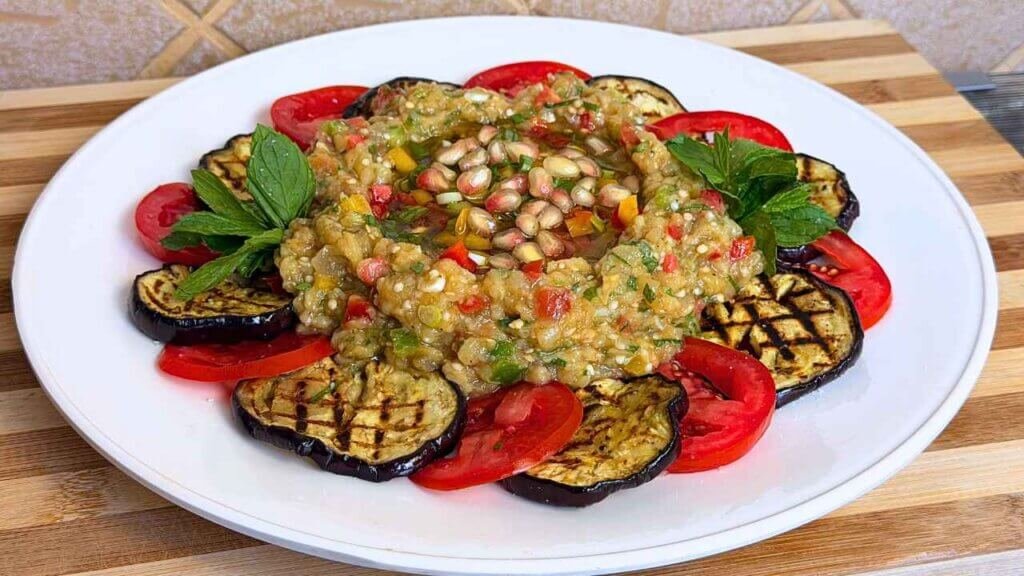 Eggplant Raheb salad with pomegranate seeds and fresh mint, surrounded by grilled eggplant and tomato slices on a white platter.