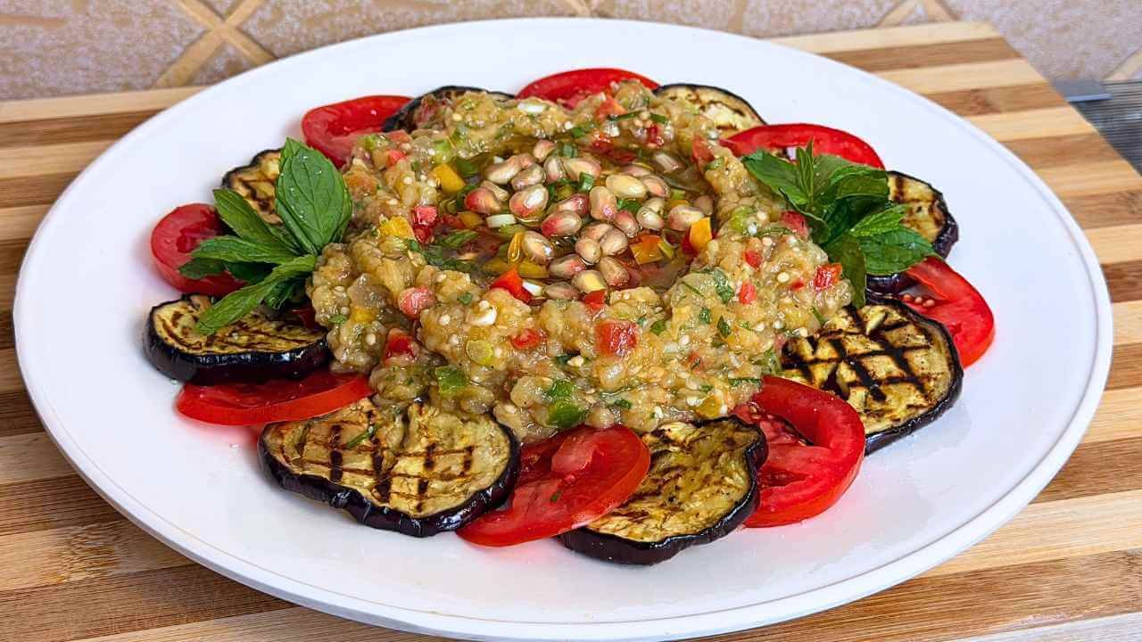 Eggplant Raheb salad with pomegranate seeds and fresh mint, surrounded by grilled eggplant and tomato slices on a white platter.