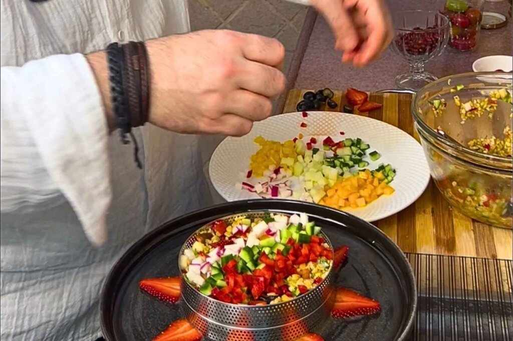 Chef François arranging colorful diced vegetables and fruits over freekeh salad inside a round plating ring on a black serving plate.