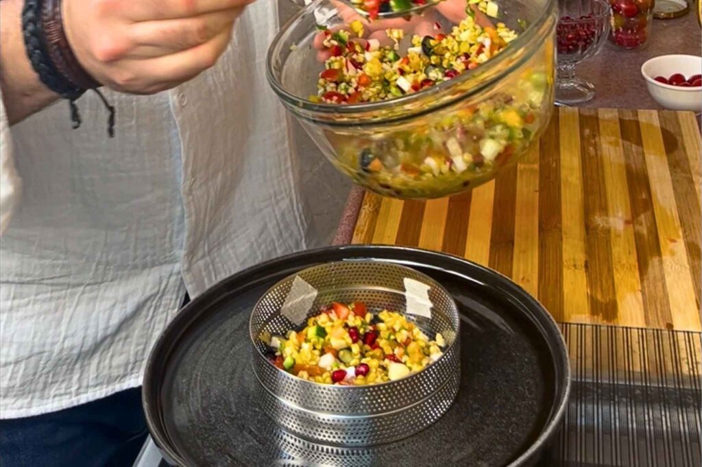 Chef François spooning colorful freekeh salad into a round stainless steel plating ring on a black serving plate.